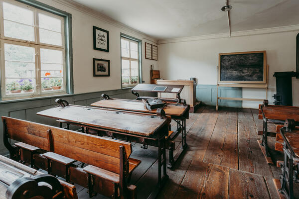 Bild vergr&ouml;&szlig;ern: helles holzvert&auml;feltes Klassenzimmer, mit alten Schulb&auml;nken auf dunklem Holzboden. Vorne befindet sich eine Tafel